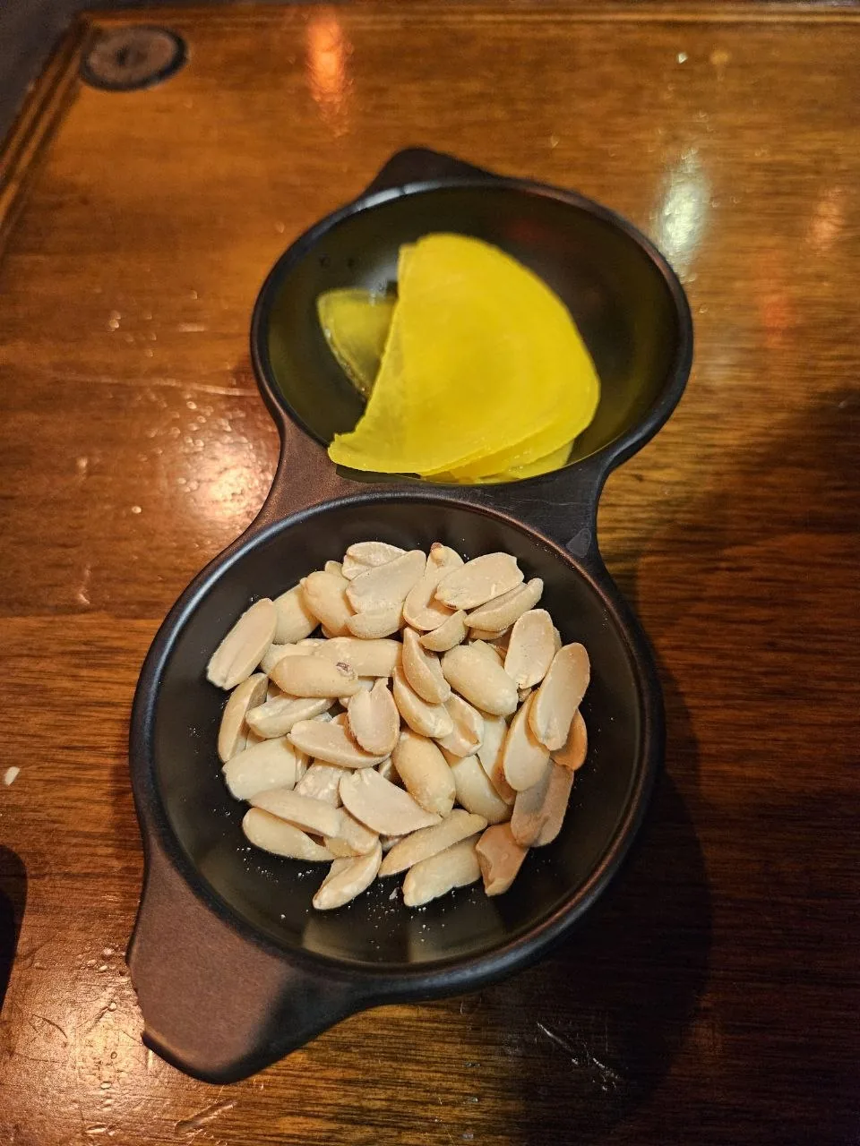 A double-compartment black ceramic dish served as a complimentary bar snack (anju), holding salted peanuts in the lower 