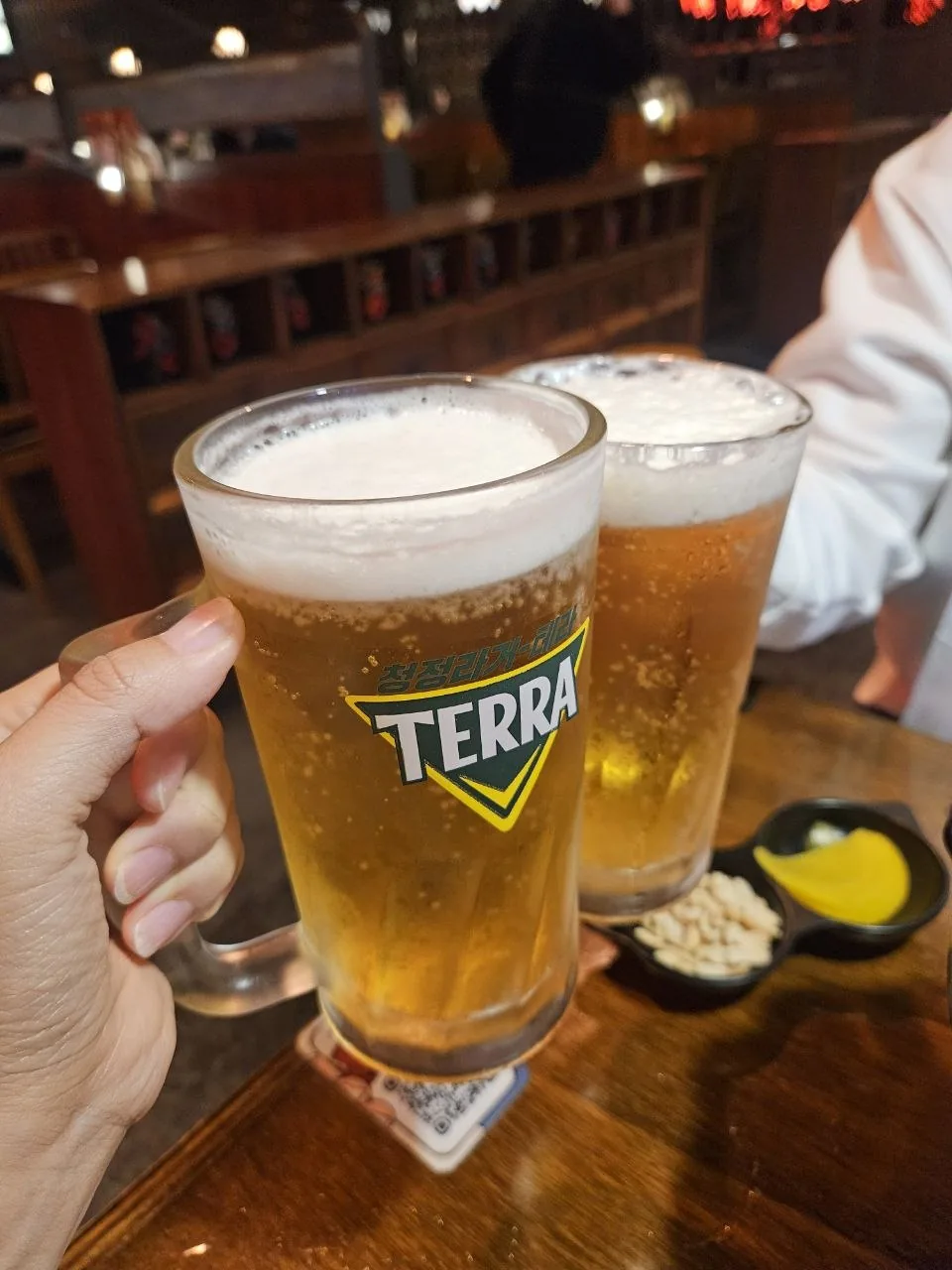 Two frosted glasses of Terra draft beer are held up at a wooden table inside a warmly lit Korean pub, with small side di