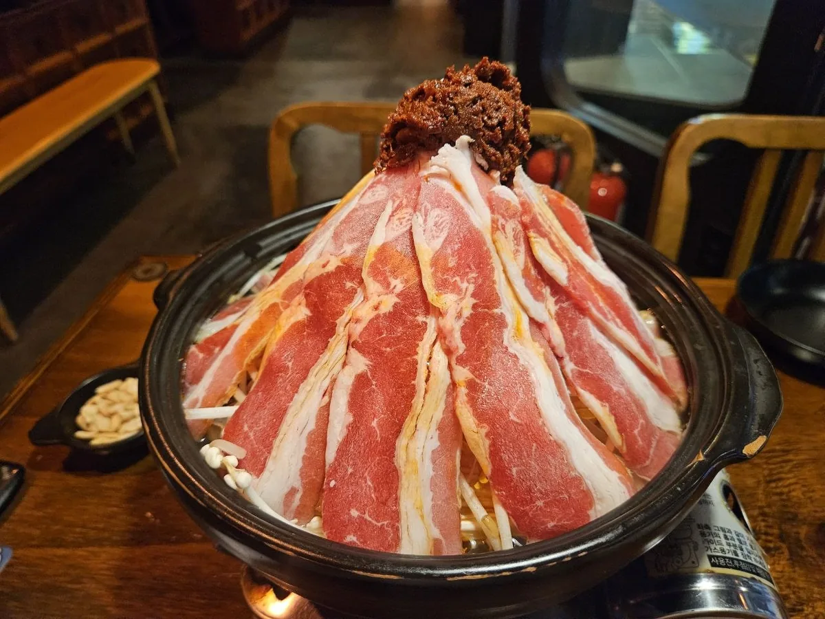 A dramatic pre-cook mala hotpot (hwasan malatang) presented in a black cast iron pot, with thick-cut pork belly slices d