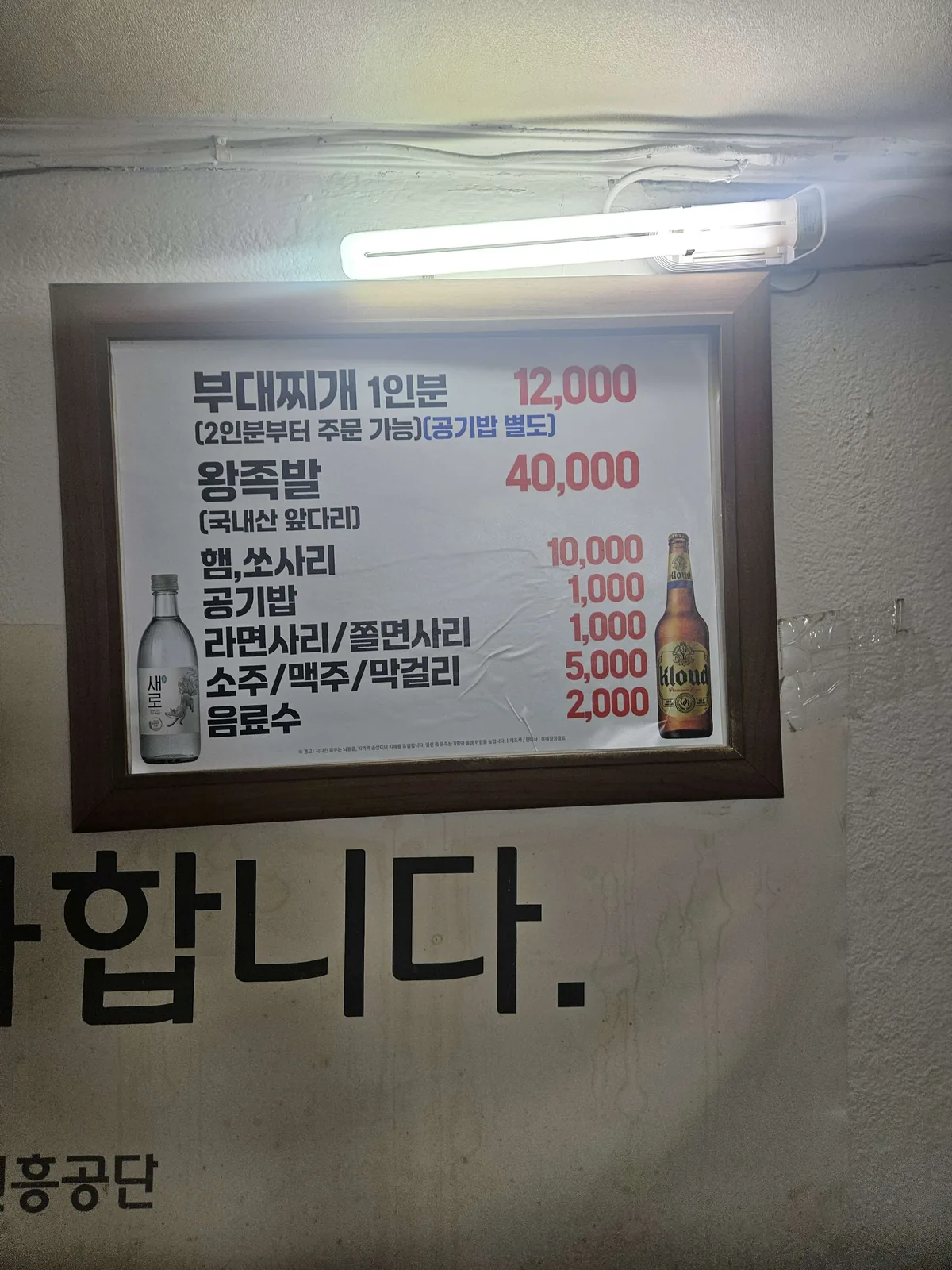 A framed printed menu board mounted on a bare wall under a fluorescent light, listing only two main dishes — budae-jjiga