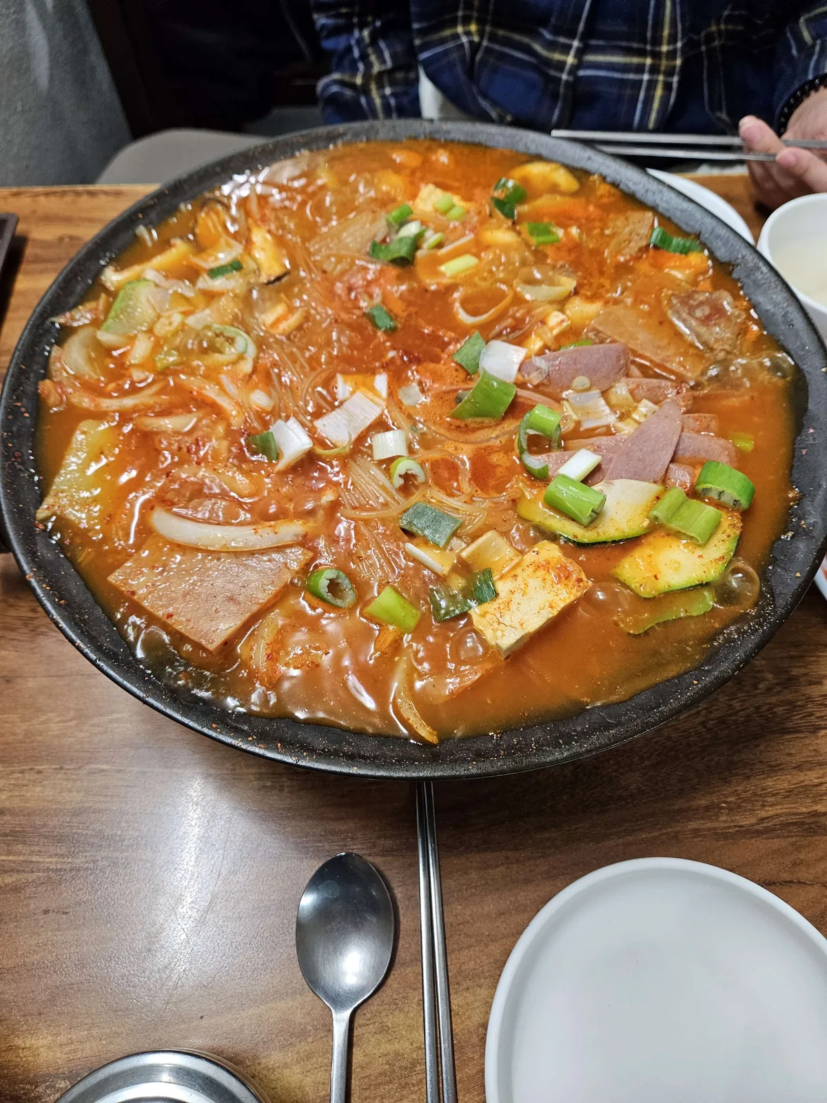 A large stone pot of bubbling budae jjigae (Korean army stew) filled with spam, tofu, kimchi, noodles, and sliced green