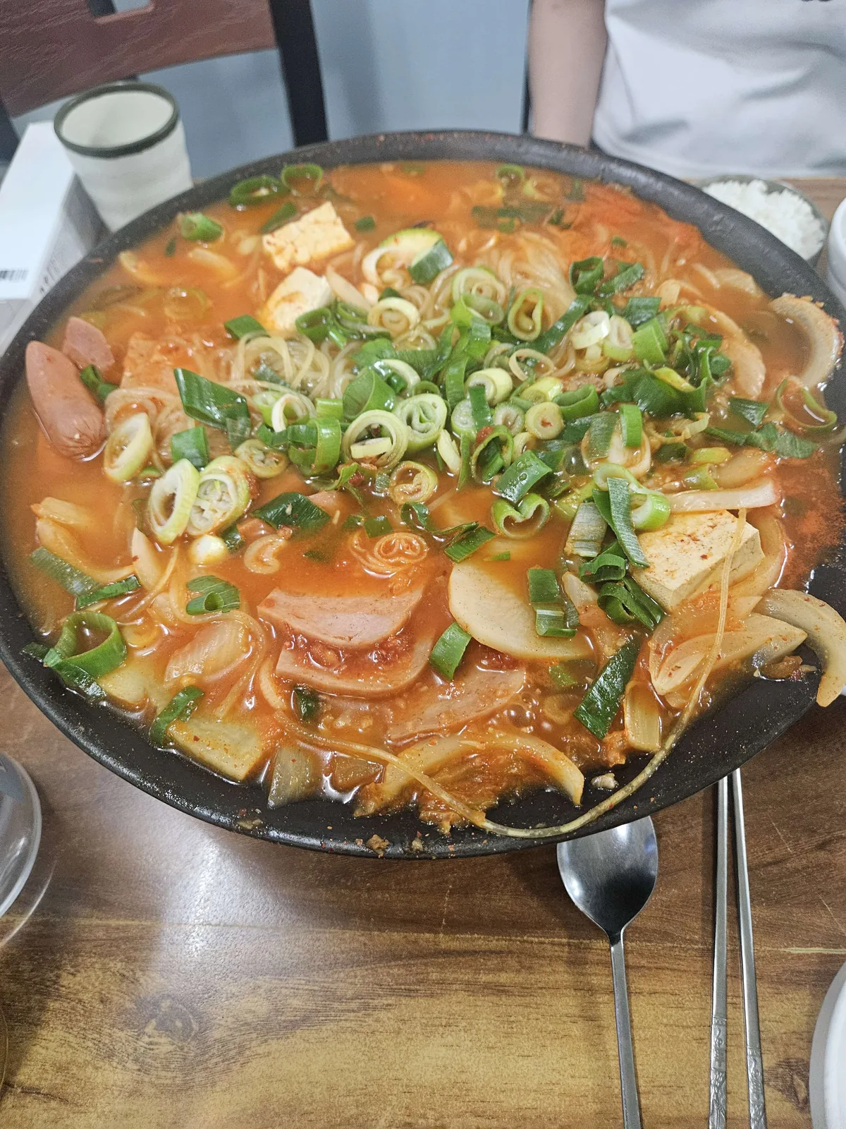 A large black cast-iron pot of budae-jjigae (Korean army stew) served at the table, packed with tofu, sausage, spam, gla