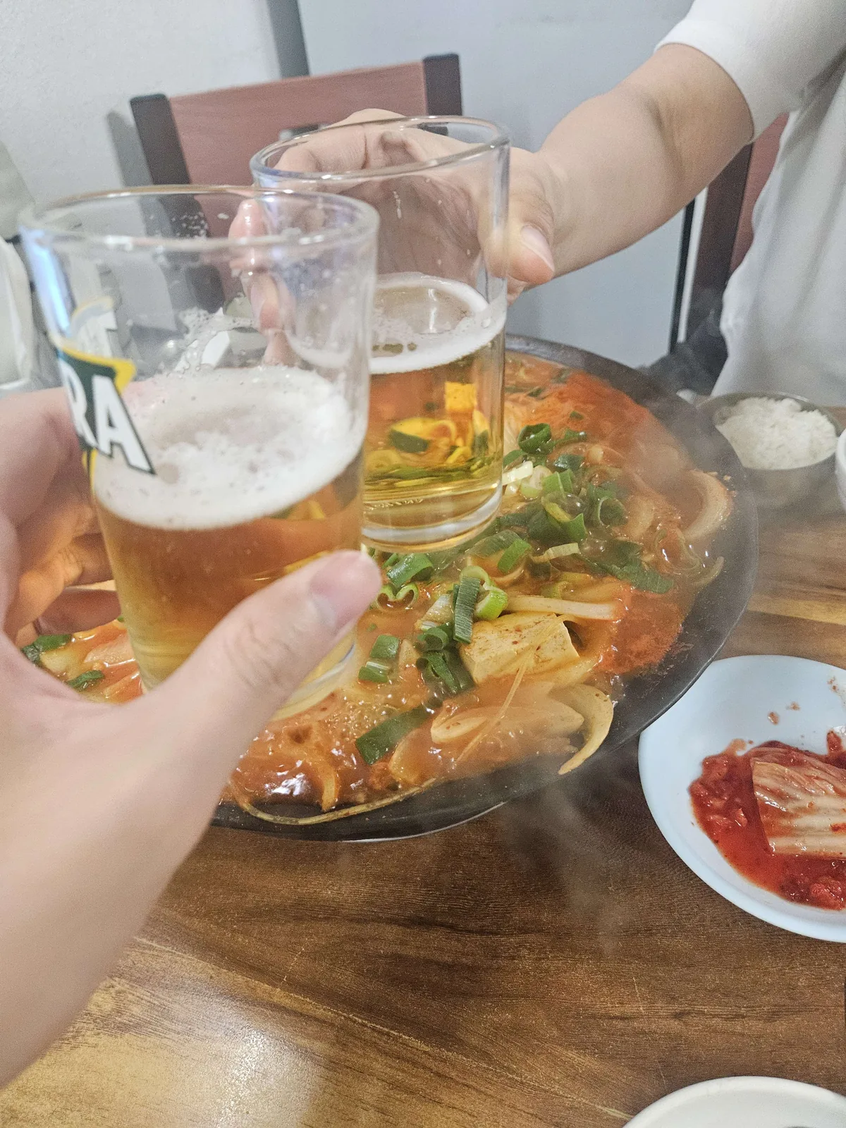 Two people clinking draft beer glasses over a bubbling pot of budae-jjigae (Korean army stew) loaded with tofu, sausage,